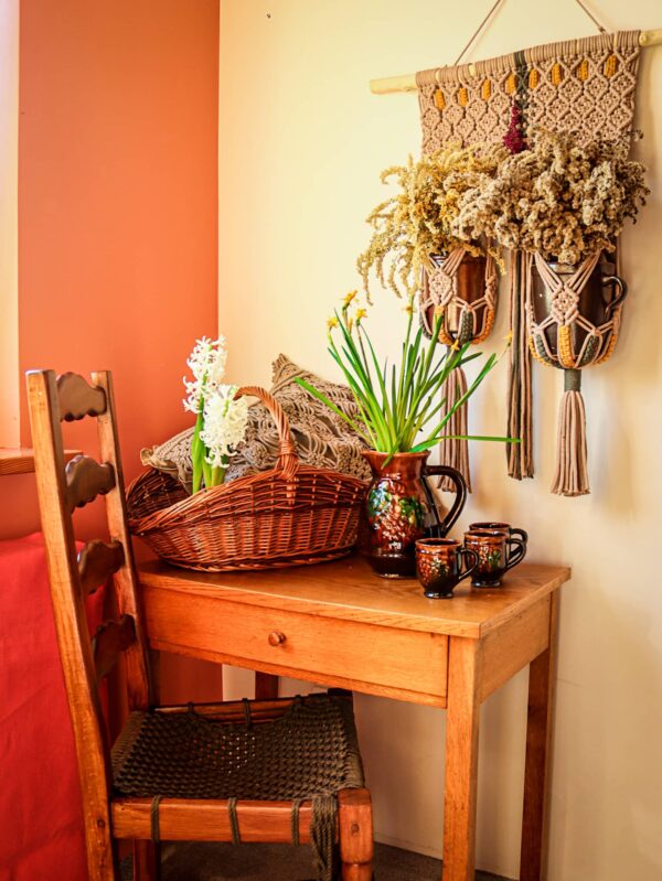Na stoliku kosz z poduszką i kwiatami, obok stoi krzesło. Nad stolikiem wisi kwietnik z dwiema donicami, a w nich suszone kwiaty