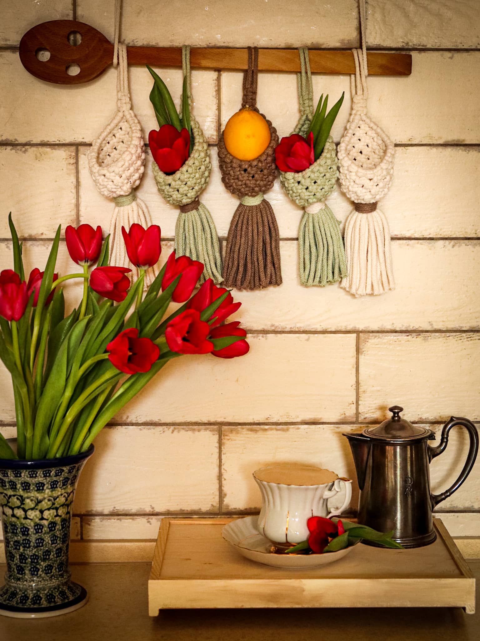 Kolorowe makramowe koszyczki wiszą na drewnianej łyżce, a w nich tulipany i cytryna
