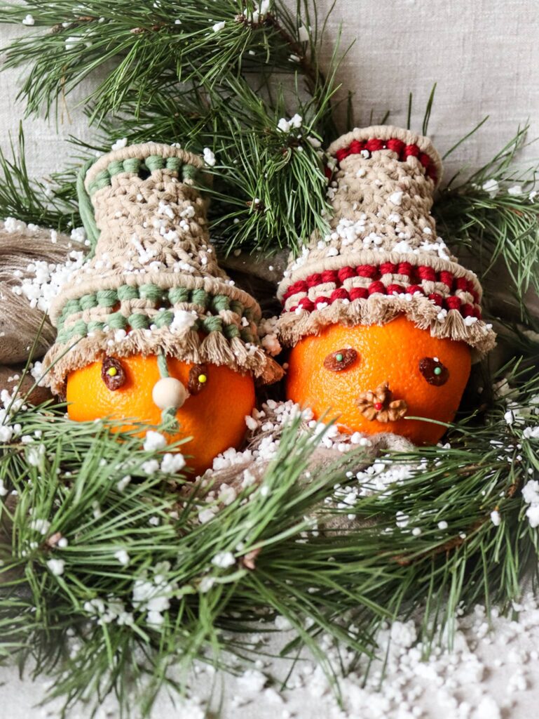 Świąteczne dekoracje - dwie pomarańcze z makramowymi dzwonkami jako czapkami wśród gałązek choinkowych i śniegu