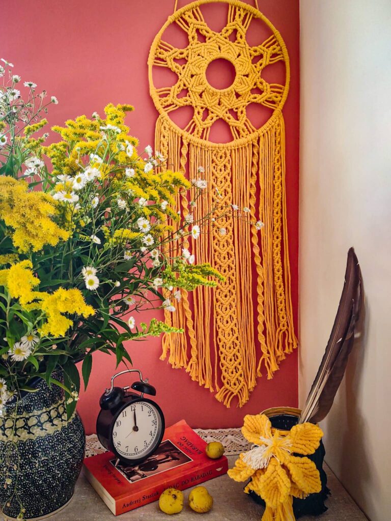 Musztardowy łapacz snów z mandalą wisi nad stolikiem, na którym stoją kwiaty, książka, budzik, wazon