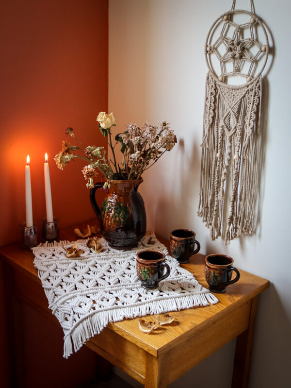 Na stoliku serweta naturalna w jodełkę, świece i dzban z suszkami, nad stolikiem beżowy łapacz snów
