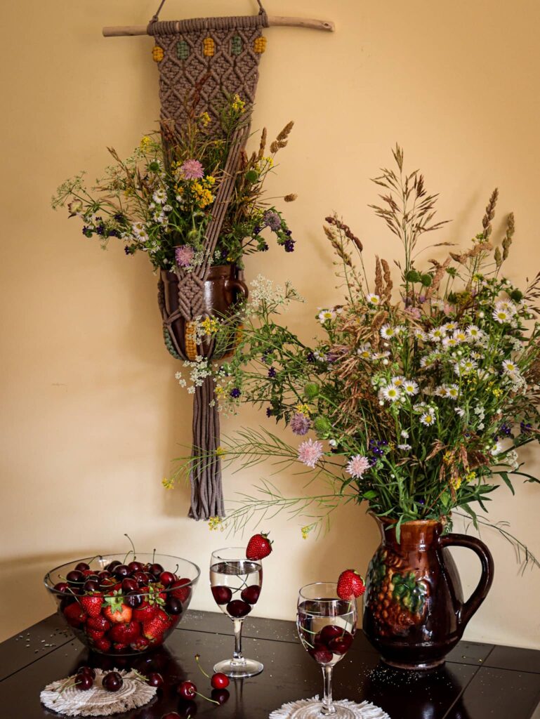Kwietnik naścienny Etno jedna doniczka wisi nad stołem, na którym wazon z polnymi kwiatami i miska z owocami