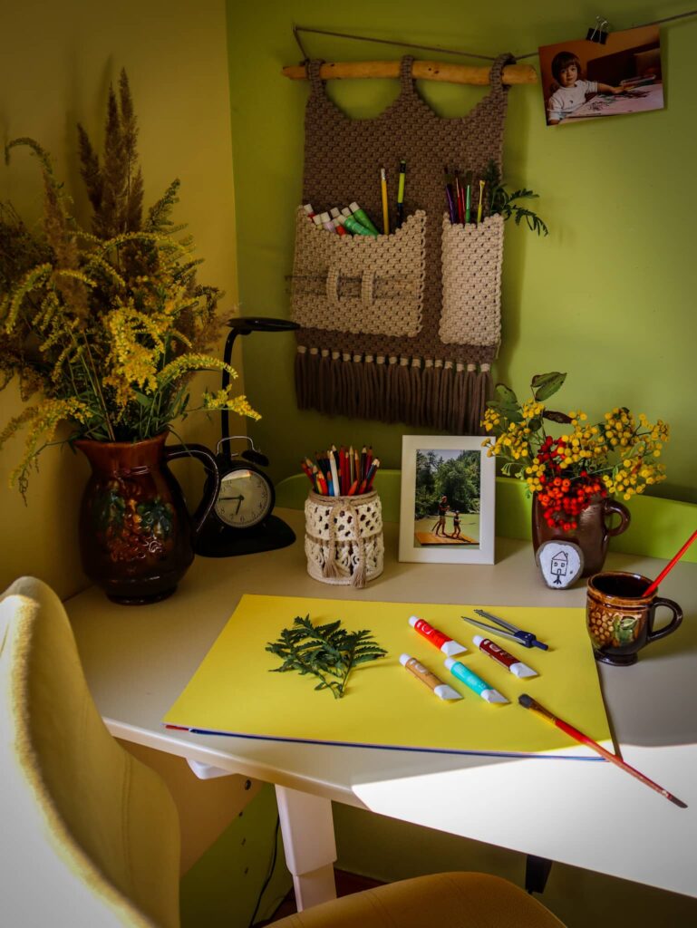 Jak dobierać kolory we wnętrzach - brązowy makramowy organizer nad białym biurkiem na tle żółtej i zielonej ściany. Na biurku malowanie