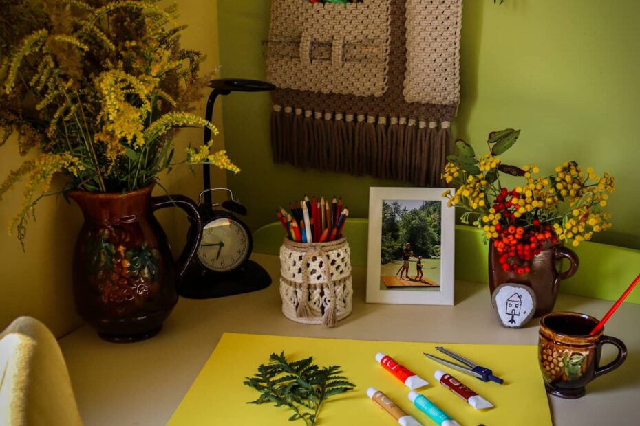 Pokrewne kolory we wnętrzach - brązowy makramowy organizer nad białym biurkiem na tle żółtej i zielonej ściany. Na biurku malowanie