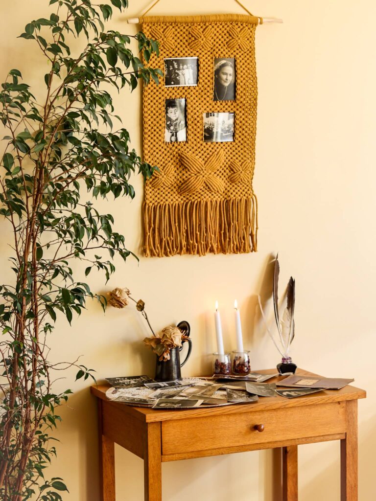 Makatka na zdjęcia wisi nad stolikiem ze starymi fotografiami, świecami i piórami. Obok duży kwiat